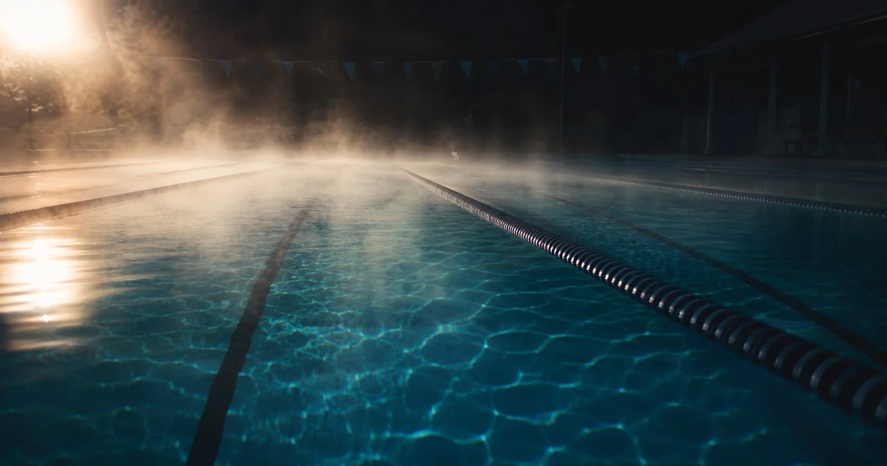 An empty swimming pool at dawn with steam rising off teal-blue water and a lane divider running into the distance